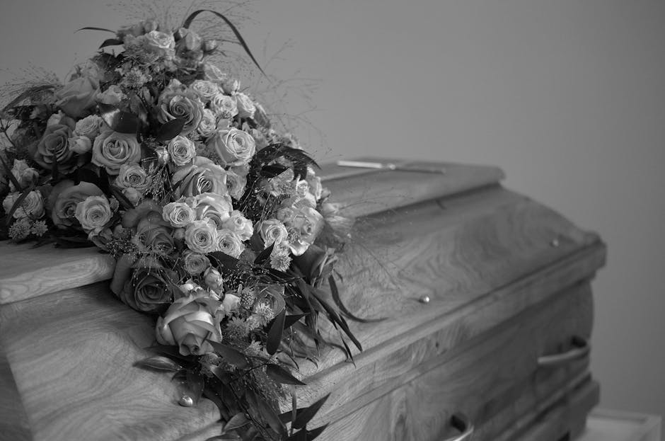 A large, delicate arrangement of fresh funeral flowers resting on the lid of a wooden casket. The bouquet includes a variety of roses in different sizes, with some fully bloomed and others as tight bu