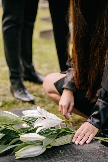 A person wearing a black dress is kneeling on the ground, arranging a floral tribute consisting of white lilies with long green leaves attached, on a dark stone surface. The bouquet is fresh, with smo
