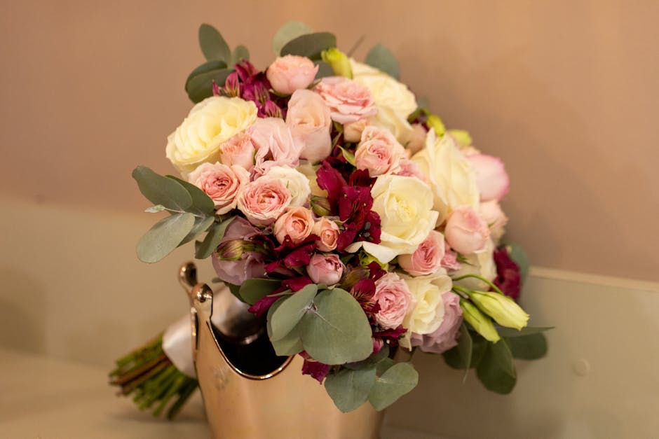 A close-up image of a fresh floral bouquet featuring a mix of pale pink and cream roses, complemented by small deep burgundy flowers and lush green eucalyptus leaves. The bouquet is arranged in a roun