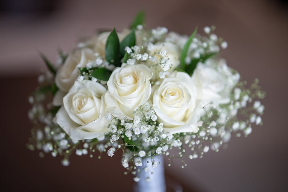 A bouquet of fresh, creamy white roses arranged with delicate sprigs of baby's breath and a few green leaves, presented in a simple, elegant style. The roses have velvety petals with a slightly open b
