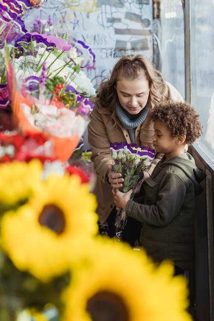A young woman and a small boy are inside a flower shop near a large window, engaged in a moment of selecting and examining a bouquet of fresh purple and white flowers. The woman, with wavy blonde hair