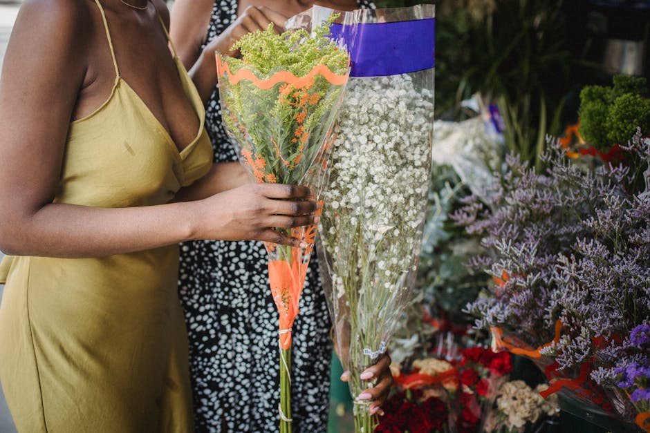 Close-up of a woman with dark skin wearing a light yellow satin slip dress, holding a tall bouquet of fresh green and white flowers wrapped in clear and orange decorative paper at a flower stand. The 